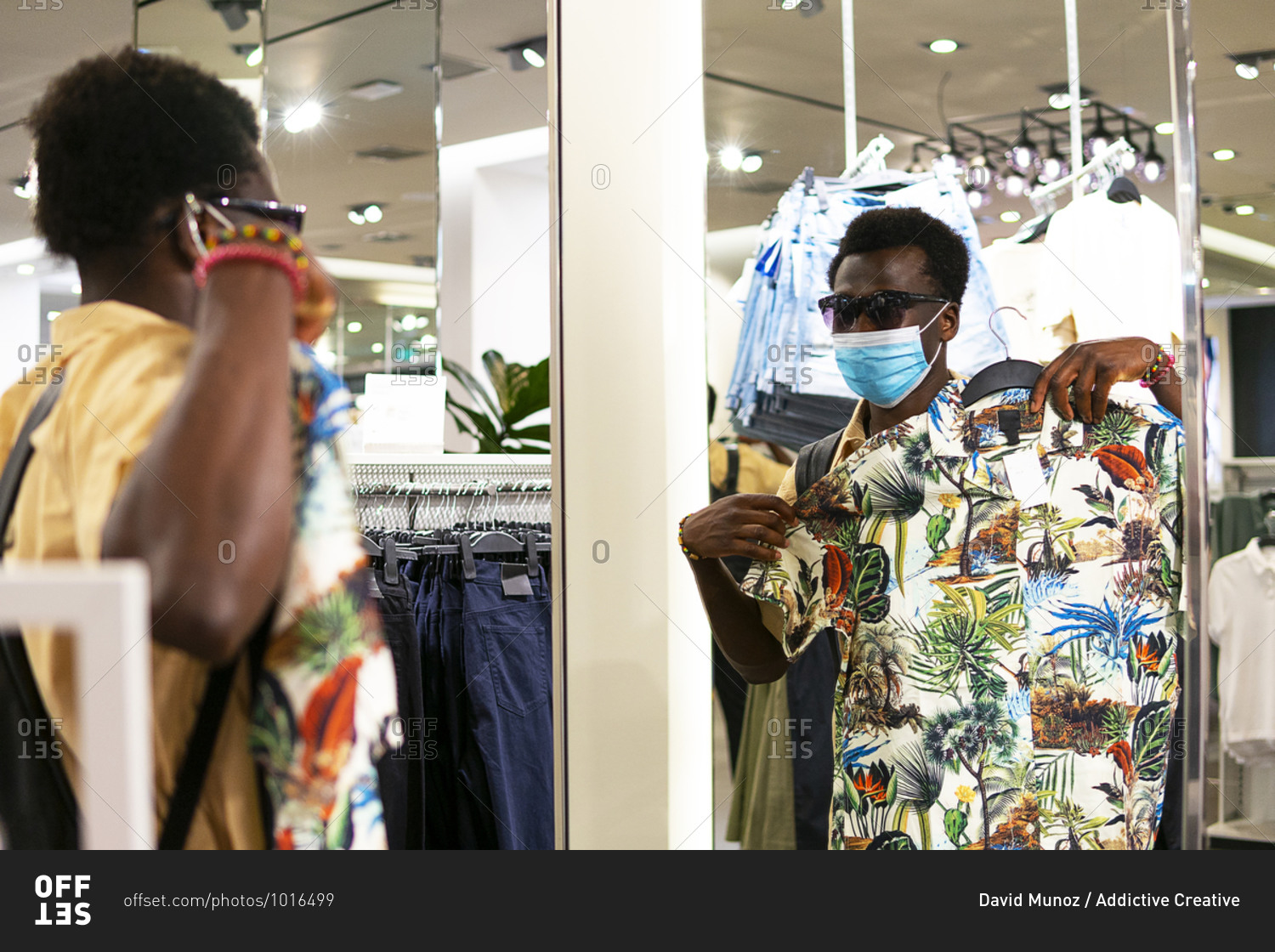 Young African American male in trendy outfit and medical mask standing in front of mirror in modern boutique and picking goods in shop during coronavirus