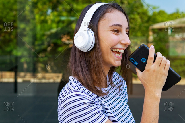 Side view of positive female listening to music in wireless headphones while standing with smartphone near glass building and looking away