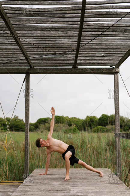 Peaceful male with naked torso standing in Vasisthasana and balancing while practicing yoga on wooden terrace and looking up