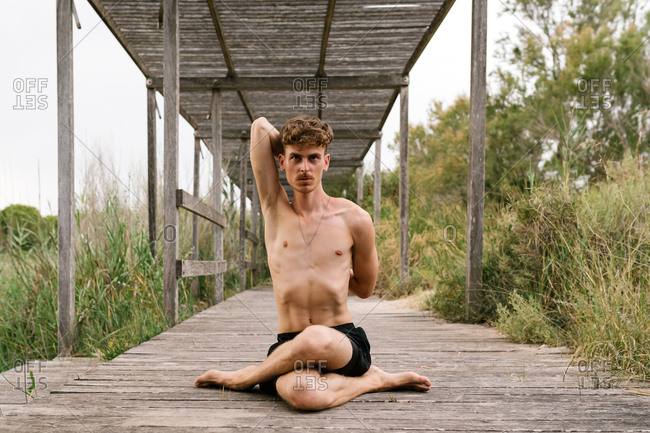 Flexible male with naked torso sitting on wooden terrace in Gomukhasana and stretching body while practicing yoga and looking forward