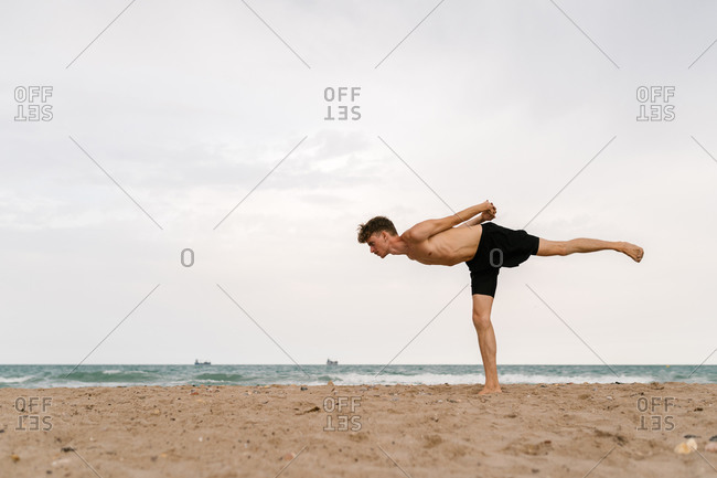 Full body side view of focused slim young male with naked torso standing in Warrior III pose with arms behind back while practicing yoga on sandy beach in cloudy summer day