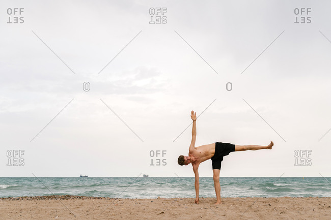 Full body side view of focused slim young male with naked torso standing in Warrior III pose with arms behind back while practicing yoga on sandy beach in cloudy summer day