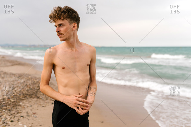 Serious slim young shirtless curly haired male with mustache and piercing looking away while standing against waving sea on beach