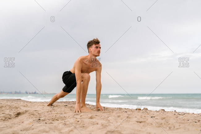 Full body of slim shirtless man doing high lunge pose during yoga practice on sandy beach near sea