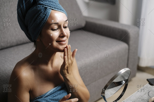 Side view of charming young female with bare shoulders and towel on head smearing facial skincare cream on face during makeup procedure at home