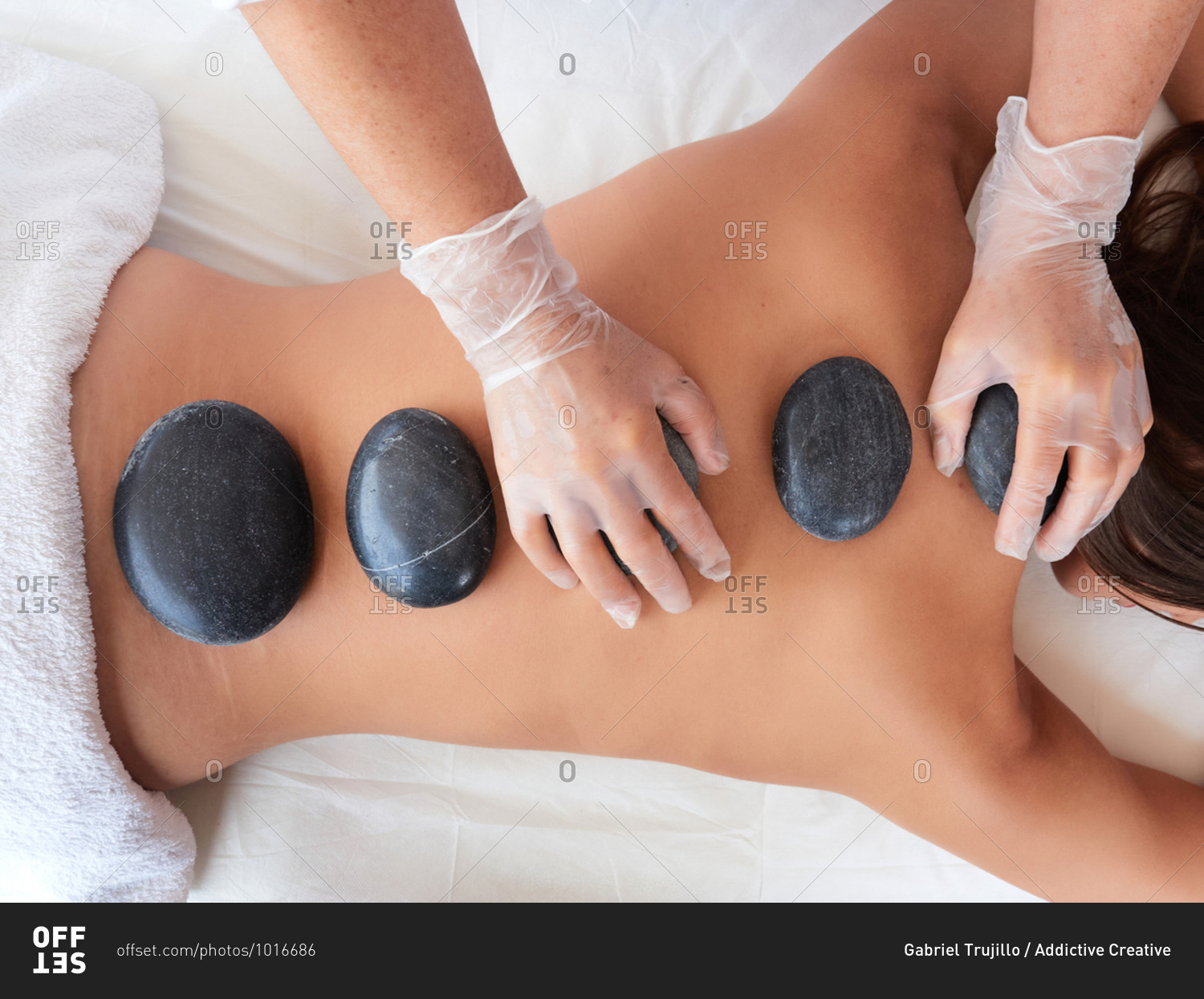 Top view of cropped unrecognizable adult artist in virus protection placing hot stones on spine of topless brunette on salon terrace