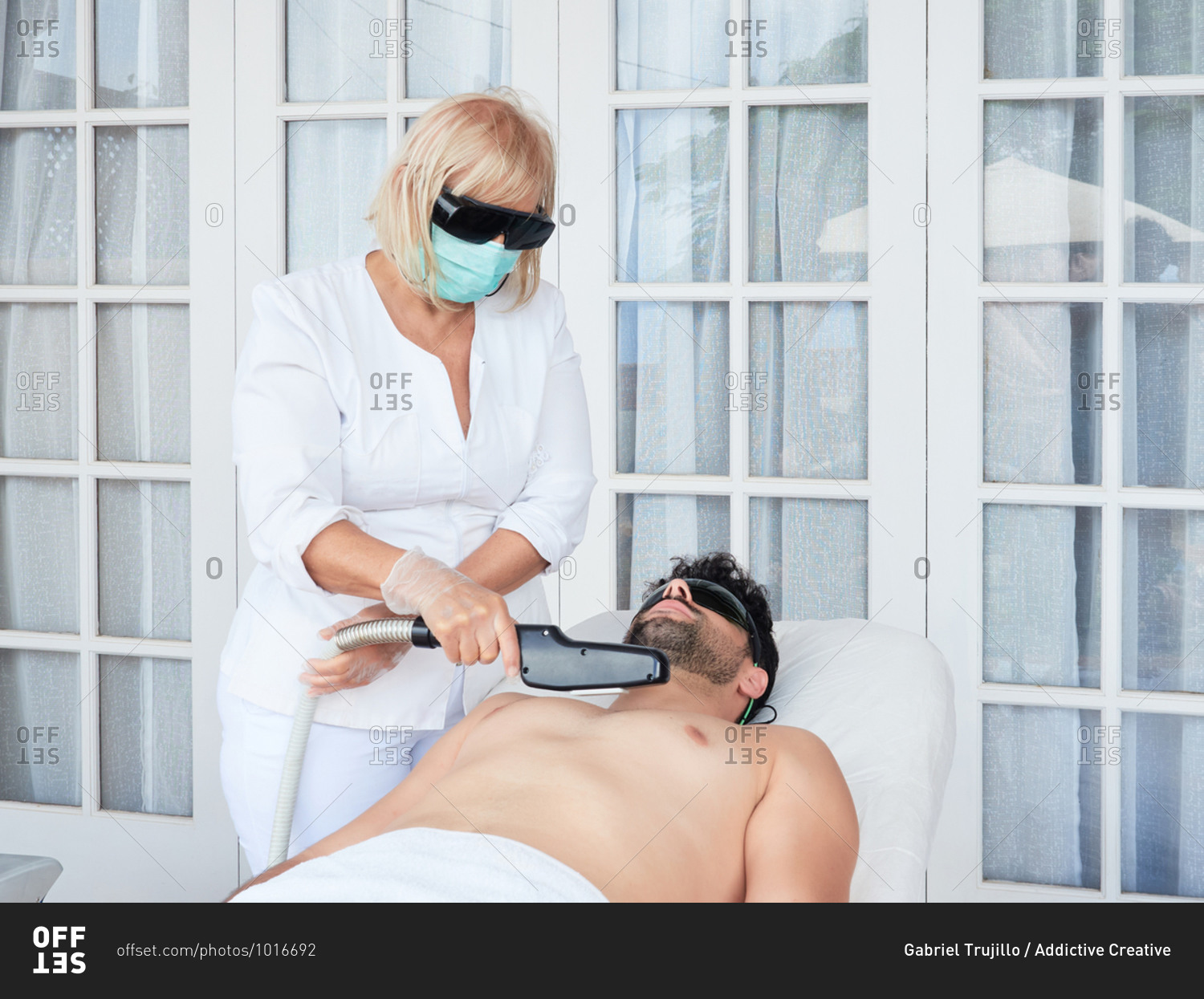 Professional woman in dark glasses and mask doing laser hair removal on chest to bearded man in salon