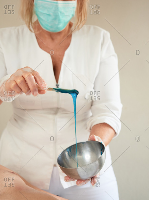 Faceless woman in medical mask and gloves stirring hot blue wax with wooden stick in bowl