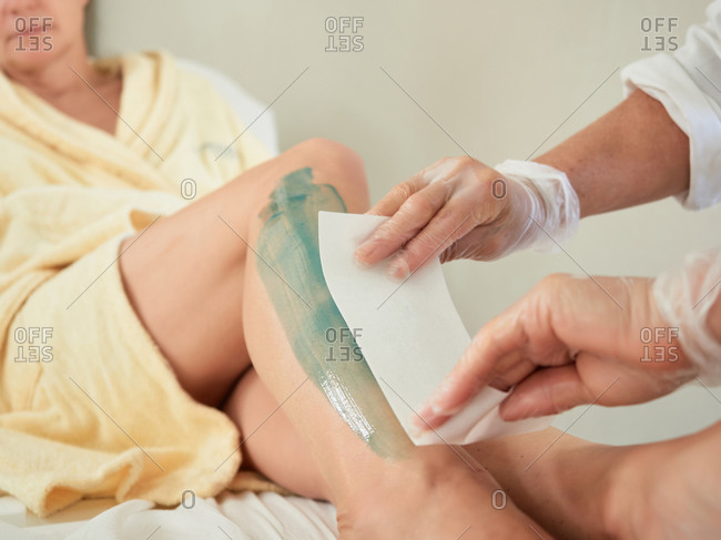 Faceless woman in gloves applying paper strip on waxed leg of client to remove hair