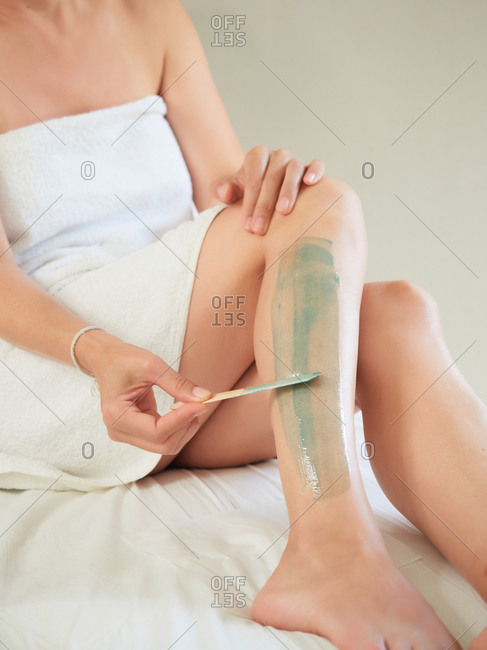 Cropped unrecognizable female removing leg hair in bathrobe using hot wax in salon