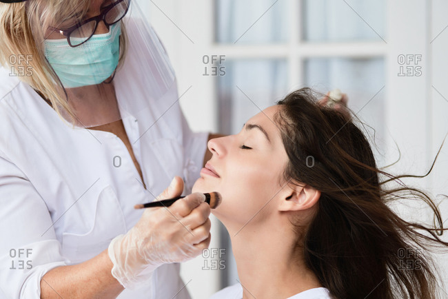 Cropped faceless professional adult woman in protective shield and mask applying foundation on face of brunette while contouring