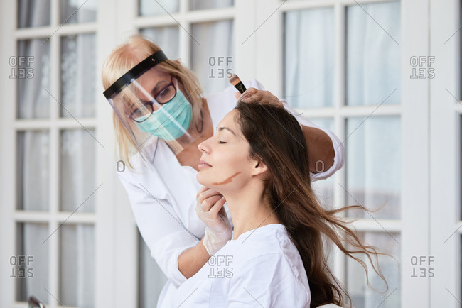 Cropped professional adult woman in protective shield and mask applying bronzer on face of brunette while contouring