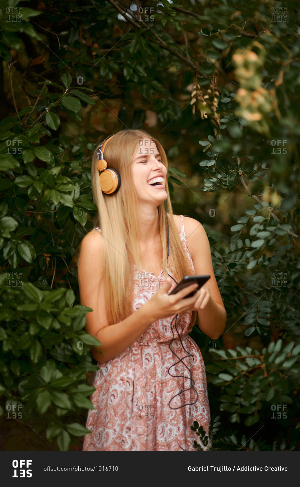 Cheerful young blond female in summer dress laughing happily while listening to music with smartphone and headphones among green foliage in garden