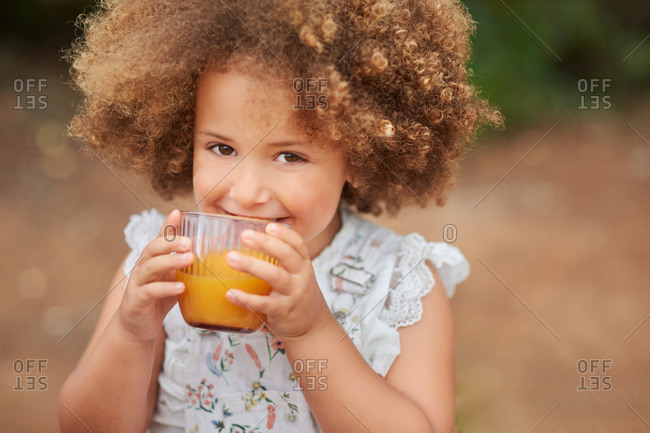 High angle of cute healthy mixed race little girl with fair Afro hair drinking fresh orange juice from glass and looking at camera against blurred nature background