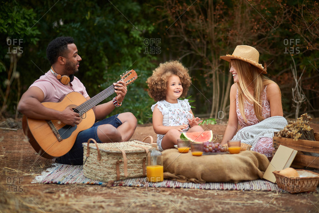 Happy young mixed race parents with cute curly haired daughter playing guitar and laughing while chilling together during picnic in summer day in green forest