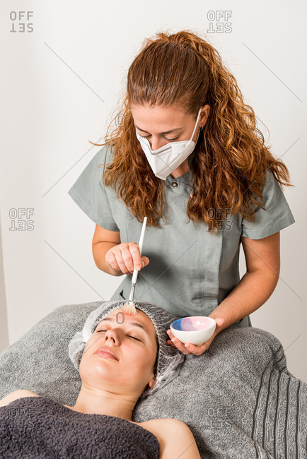 Young female beauty specialist in protective mask and uniform spreading mask on face of relaxed client while working in modern salon