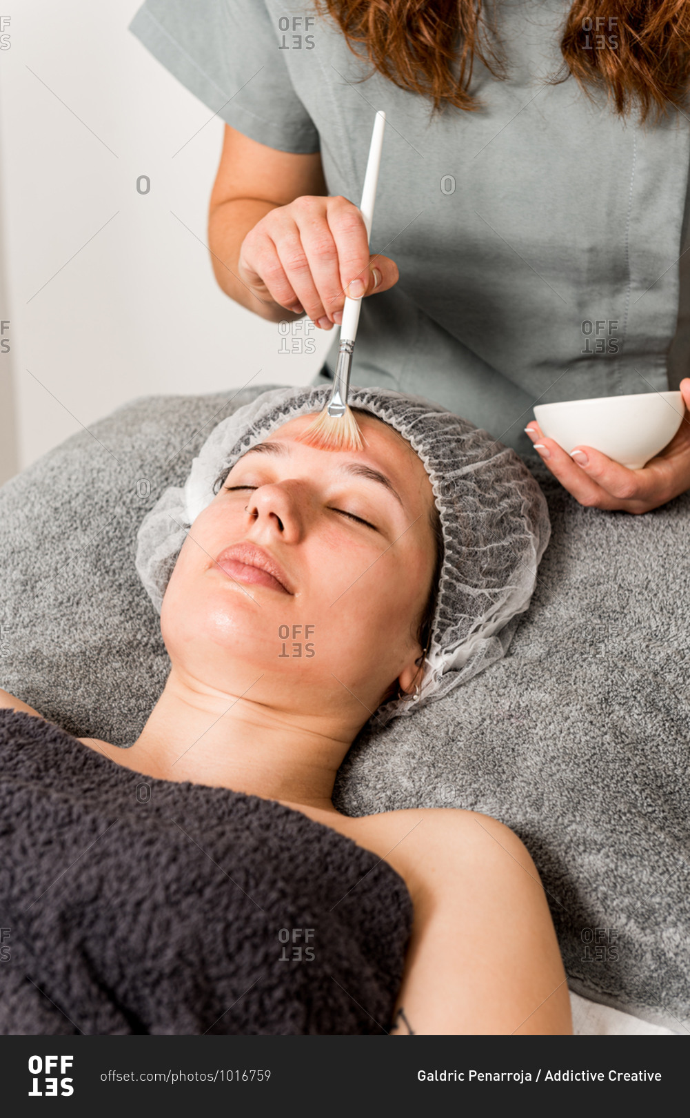 Cropped unrecognizable female beauty specialist in uniform spreading mask on face of relaxed client while working in modern salon