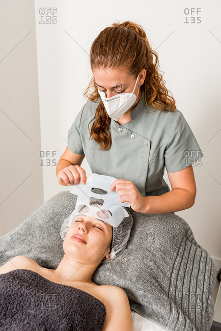Beautician in protective mask removing moisturizing cloth mask from face of relaxed female client during professional treatment in modern clinic