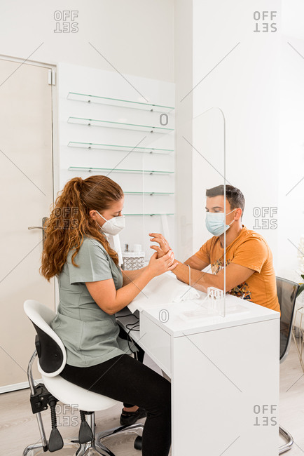 Side view of young male in protective mask getting professional manicure procedure from female specialist in modern beauty center