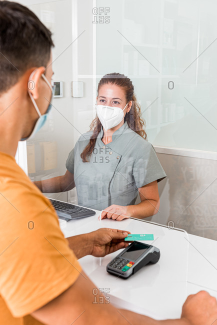 Young female specialist in protective mask holding card reader while male patient paying for treatment with credit card in modern clinic