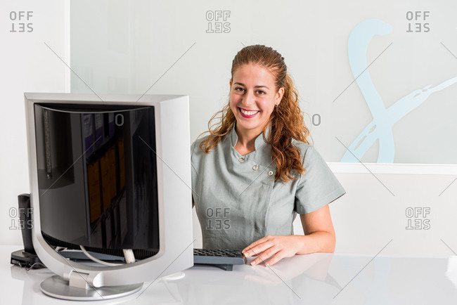 Pleasant smiling young female receptionist in uniform checking information on computer while sitting at desk in contemporary clinic office