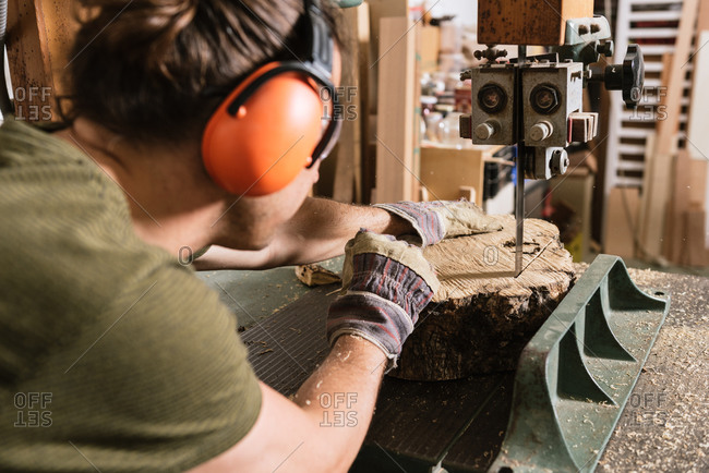 Male carpenter in protective ear muffs and goggles cutting piece of wood with band saw in shabby workshop