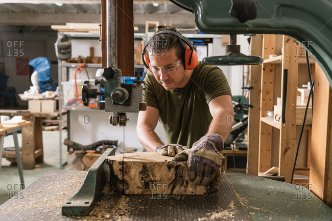 Focused male carpenter in protective ear muffs and goggles cutting piece of wood with band saw in shabby workshop