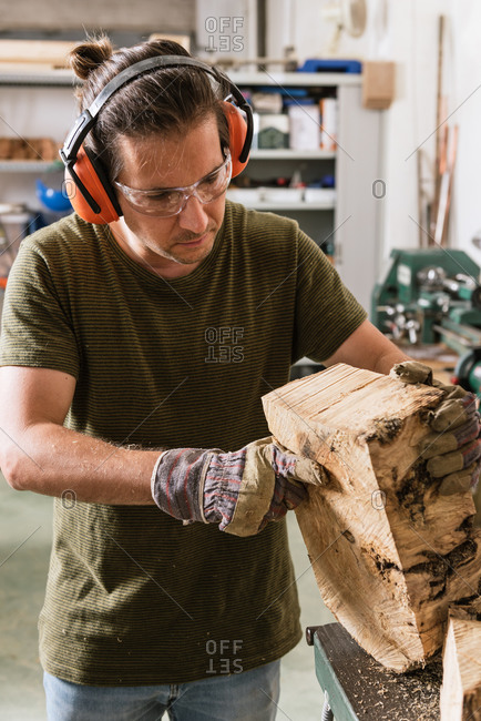 Concentrated male woodworker in protective goggles and industrial ear muffs standing at workbench with wood