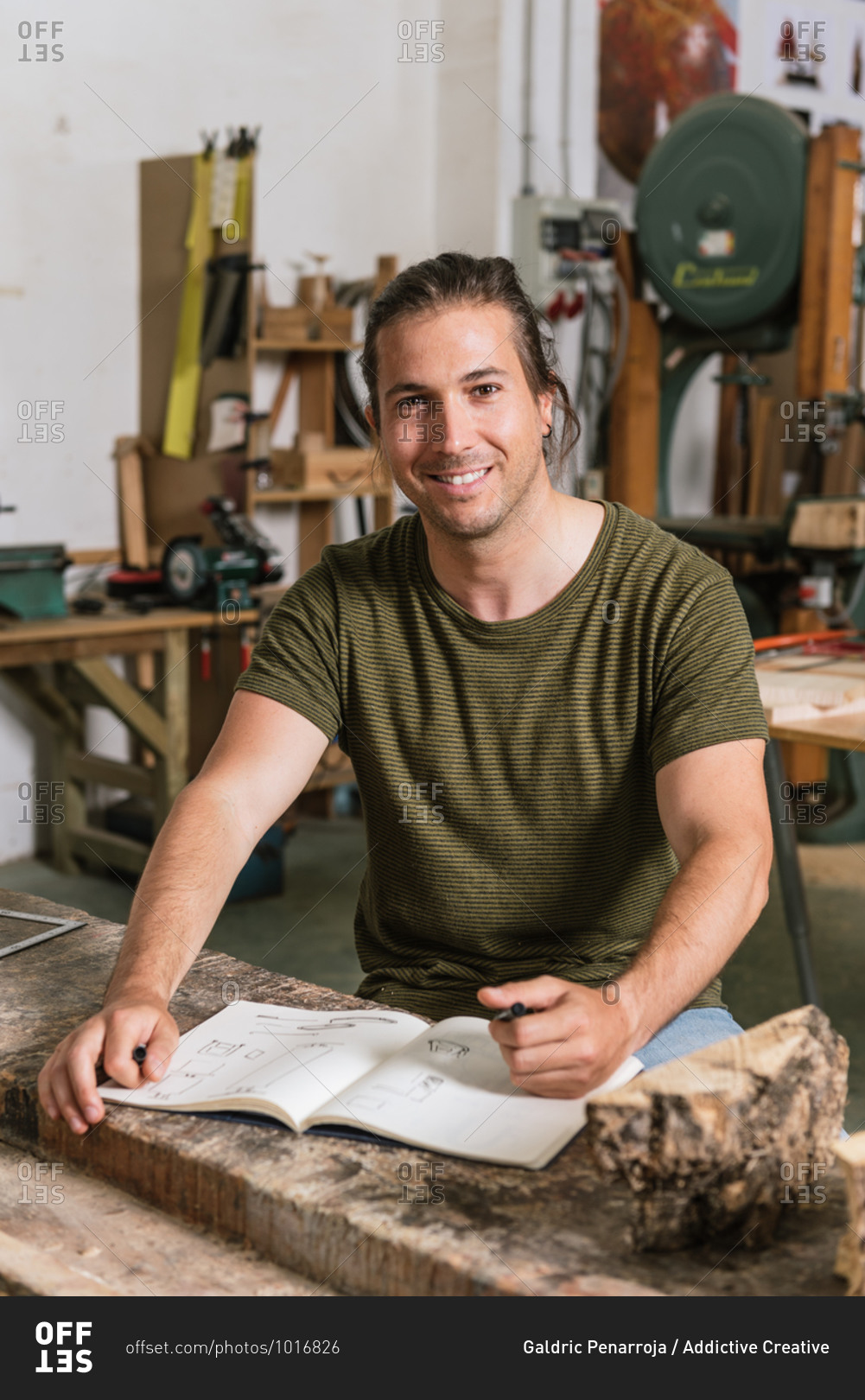 Cheerful male carpenter standing at workbench and taking notes in notebook while working in garage