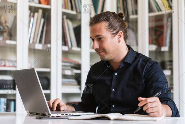 Focused businessman in formal shirt sitting at table while doing paperwork and typing on notebook in bright workspace