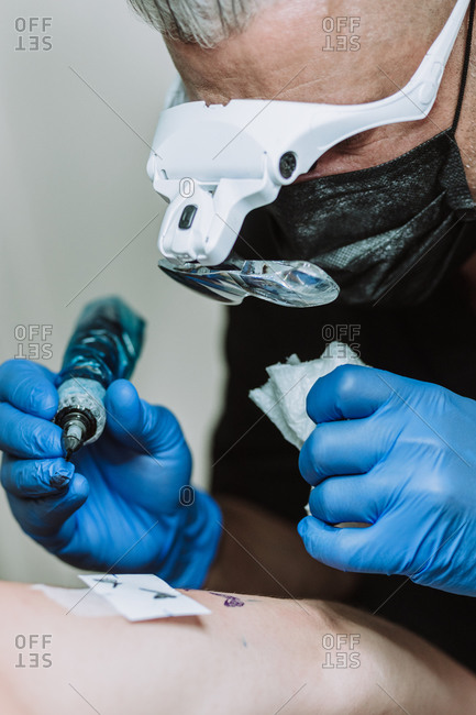 Busy male tattoo artist in gloves and mask using machine with ink and creating picture on arm of client in salon