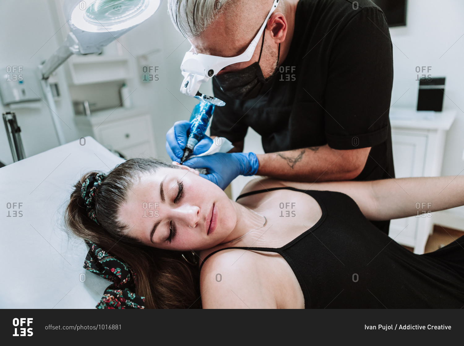 Side view of mature male tattooist in protective mask and medical glasses with tattoo machine making micro tattoo on ear of female customer