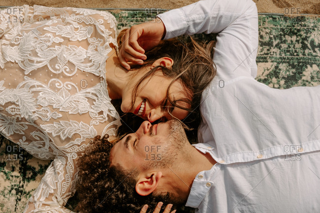 From above romantic newlywed young couple in stylish clothes lying opposite with face to face with eyes closed cuddling on vintage rug on sandy beach