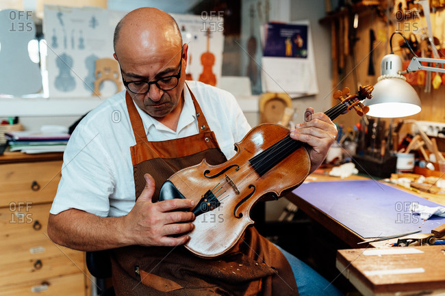 Mature male luthier in apron and glasses sitting on chair and holding restored violin while working in workshop
