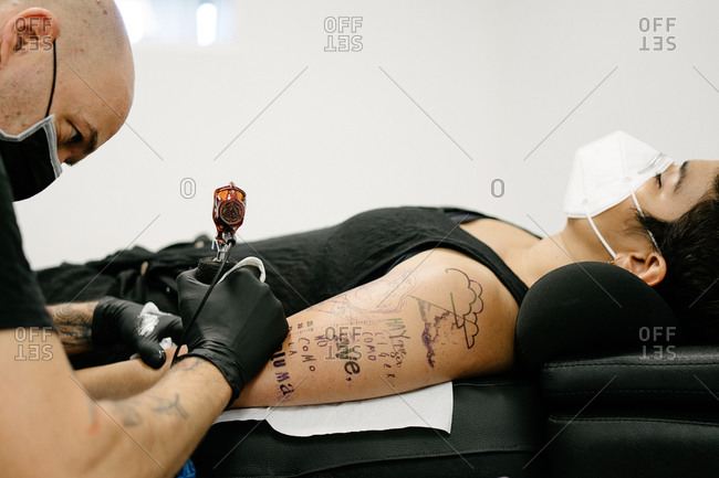 Crop male tattooist with machine making tattoo on arm of woman lying on table with closed eyes in salon