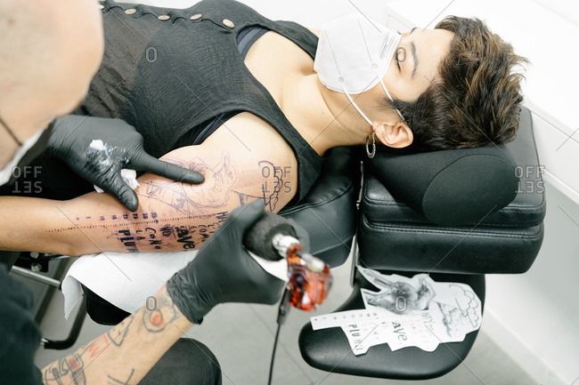 Crop male tattooist with machine making tattoo on arm of woman lying on table in salon
