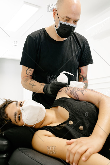 Male tattooist in medical mask treating tattoo on arm of female customer with special liquid in contemporary salon