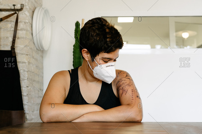 Delighted female in protective mask sitting at table in modern salon after procedure and enjoying new tattoo on arm while looking down