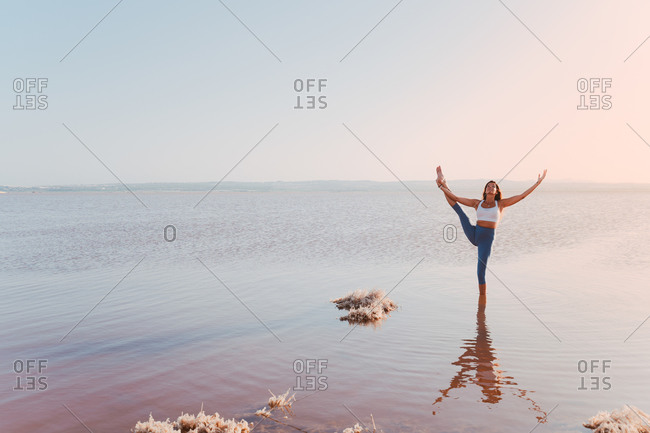 Full body of female in sportswear standing in lake water and performing standing split asana with arm raised while practicing yoga in sunny evening