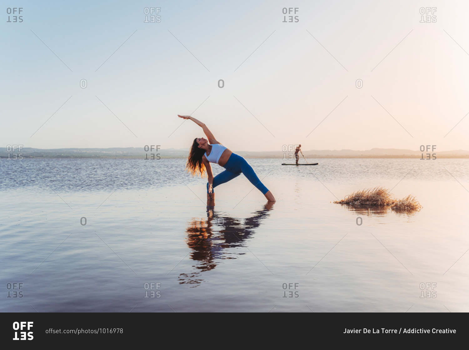 Full body of unrecognizable slim female in sportswear stretching in Extended Side Angle yoga asana during practice on lake shore against cloudless sunset sky with distant man surfing on paddle board in background
