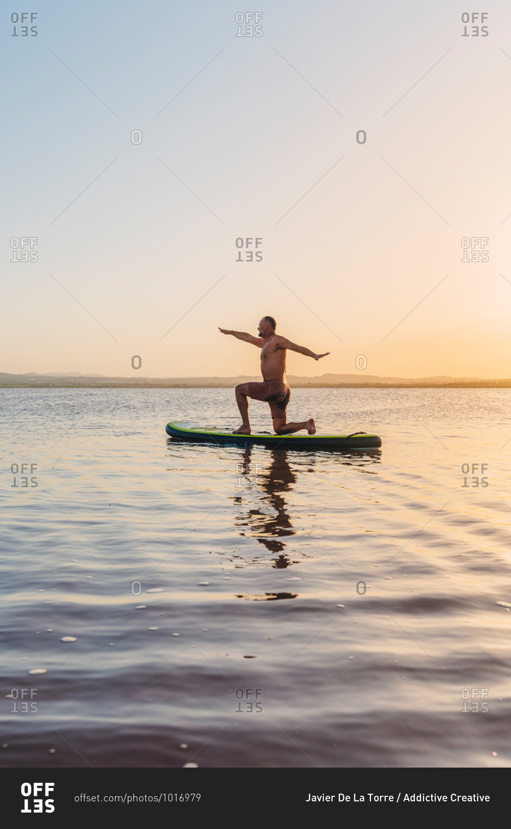 Full body side view of unrecognizable man doing variation of Crescent Lunge on Knee asana while floating on paddle board in calm lake water during sunset