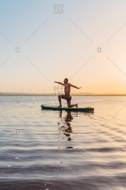 Full body side view of unrecognizable man doing variation of Crescent Lunge on Knee asana while floating on paddle board in calm lake water during sunset
