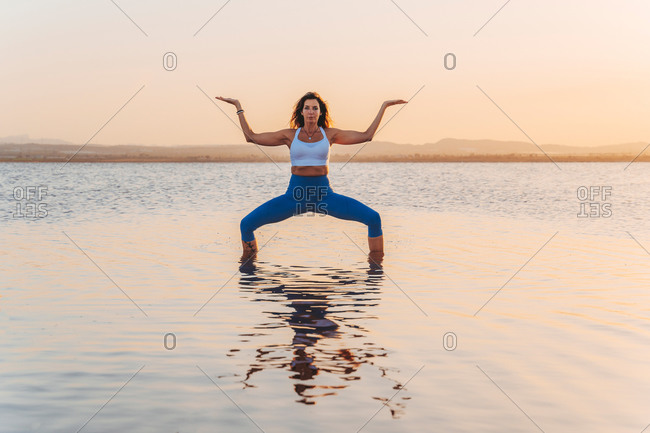 Full length of young slim female in activewear standing in Goddess yoga asana in calm lake water during summer sunset