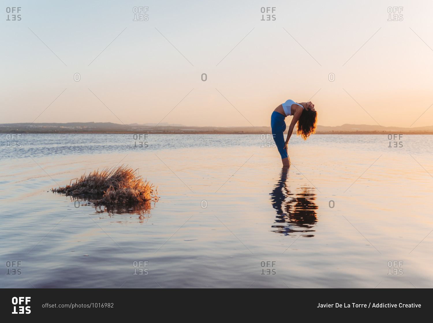 Full length side view of young flexible female in sportswear practicing yoga and doing Standing Backward pose with eyes closed in calm lake water during sunset