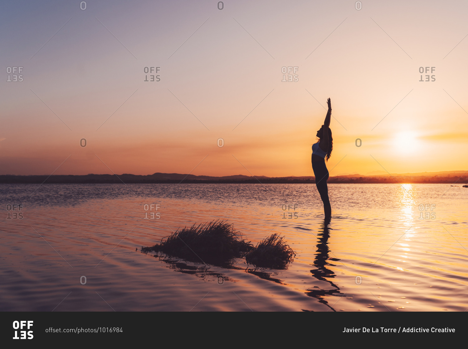 Full body side view of unrecognizable slim female in activewear standing in Upward Salute Pose with arms up in lake water while practicing yoga during sunset