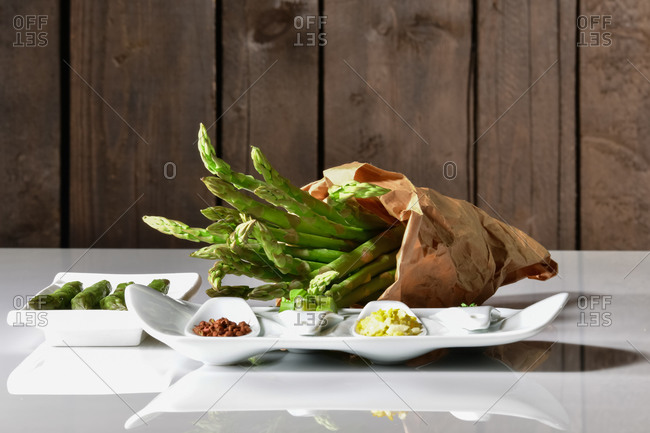 Paper bag filled with fresh asparagus surrounded by plates with more asparagus and assorted vegetables on a table. Healthy food concept.