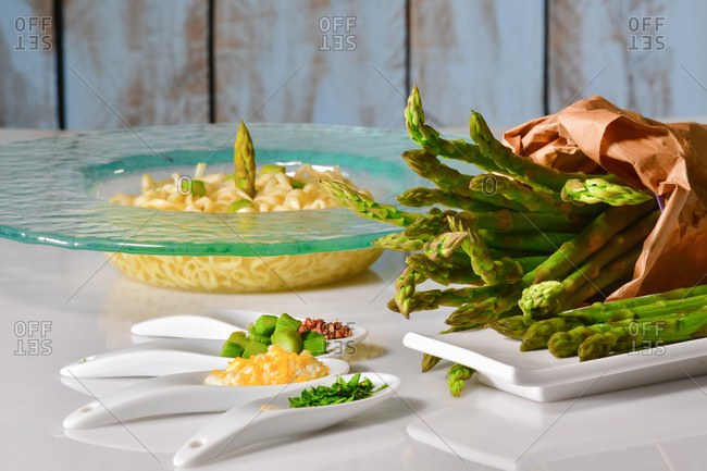 Close up of an assortment of plates with chopped vegetables and asparagus, with a paper bag filled with asparagus and a pasta dish at the back. Healthy food concept.