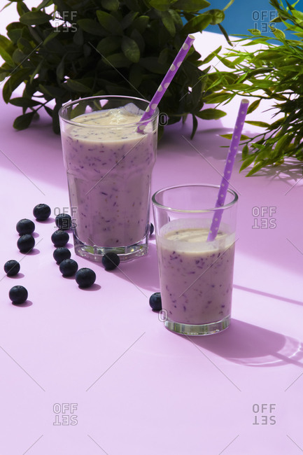Tasty healthy blueberry smoothie served in glasses with straws on table with fresh berries and green plants on purple surface