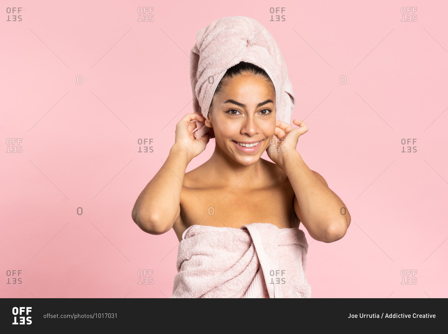 Happy young ethnic bare shouldered female with bath towel on head and perfect olive skin looking at camera against pink background while representing beauty and skincare concept