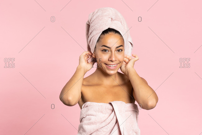 Happy young ethnic bare shouldered female with bath towel on head and perfect olive skin looking at camera against pink background while representing beauty and skincare concept